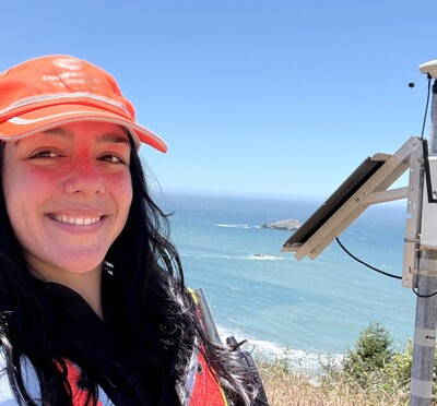Heather Maran stands beside a coastal monitoring instrument overlooking the Oregon Coast with vegetation and ocean behind her.