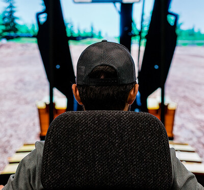 A rear view of a person seated in a heavy equipment simulator, facing a large screen that displays a virtual construction site.