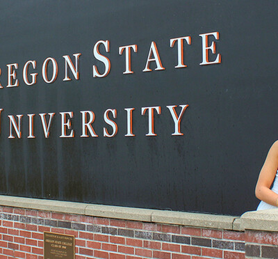 Megan Thomas wears a graduation cap and stole and stands outdoors leaning against a large black Oregon State University sign.