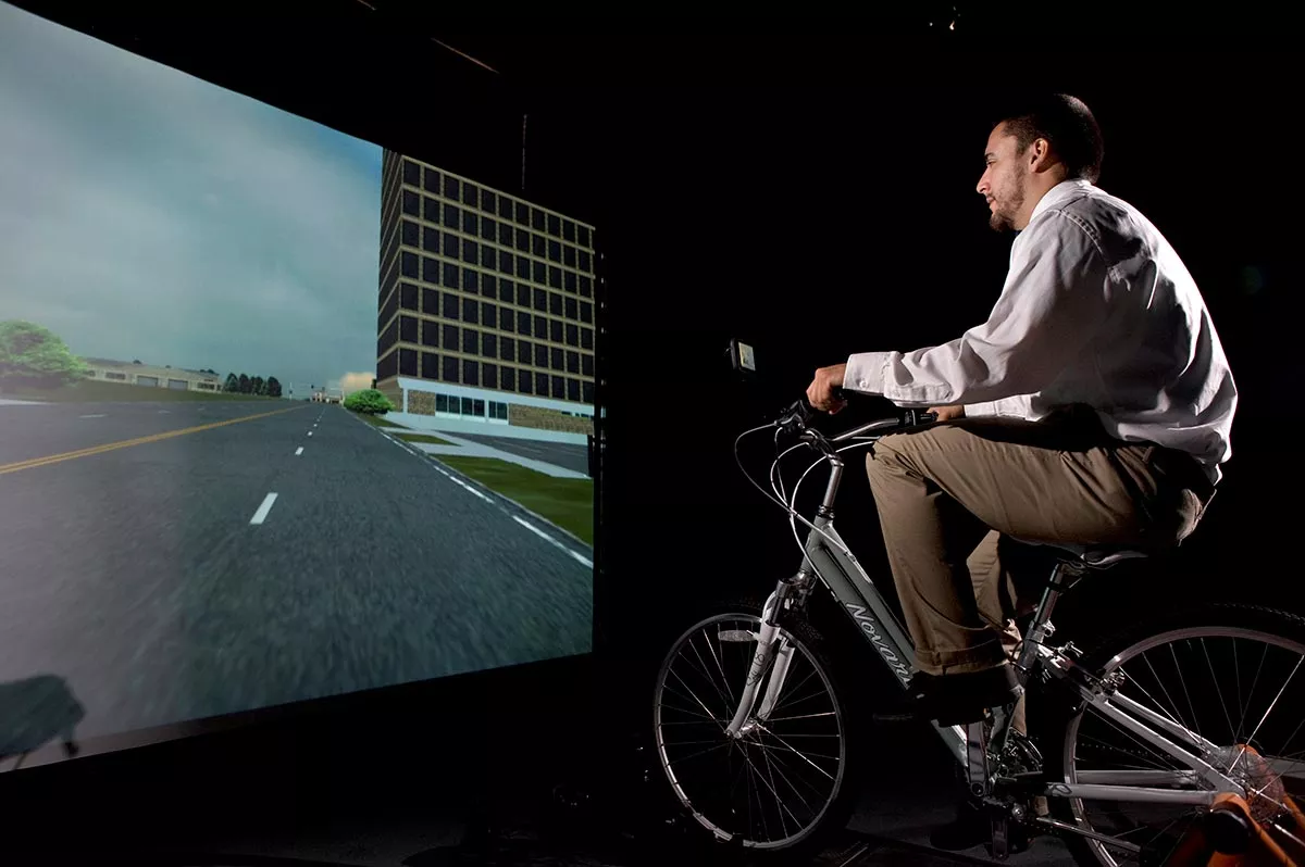 A person riding a bicycle in front of a screen with a simulation of a road.