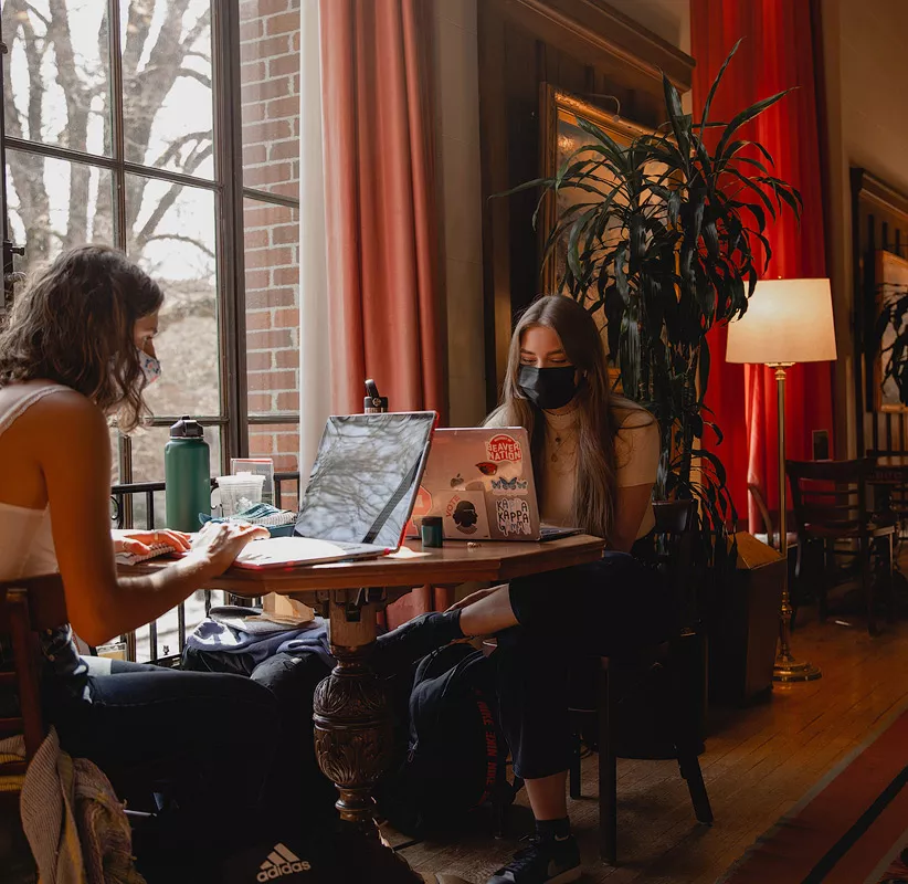 2 Person sitting near to each other, working on their laptops near a window.