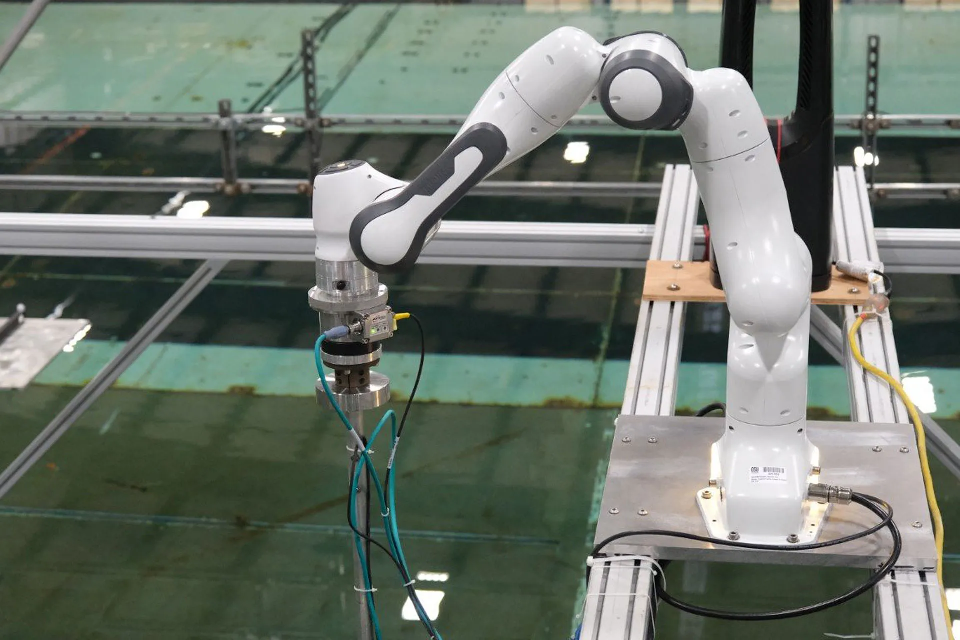 A robotic arm sitting on two supports over an indoor water testing area. Caption: Oregon State researchers delivered simulated wind movements to a wave energy device by programming a robotic arm to push and pull the mast of a 1/50 scale device.