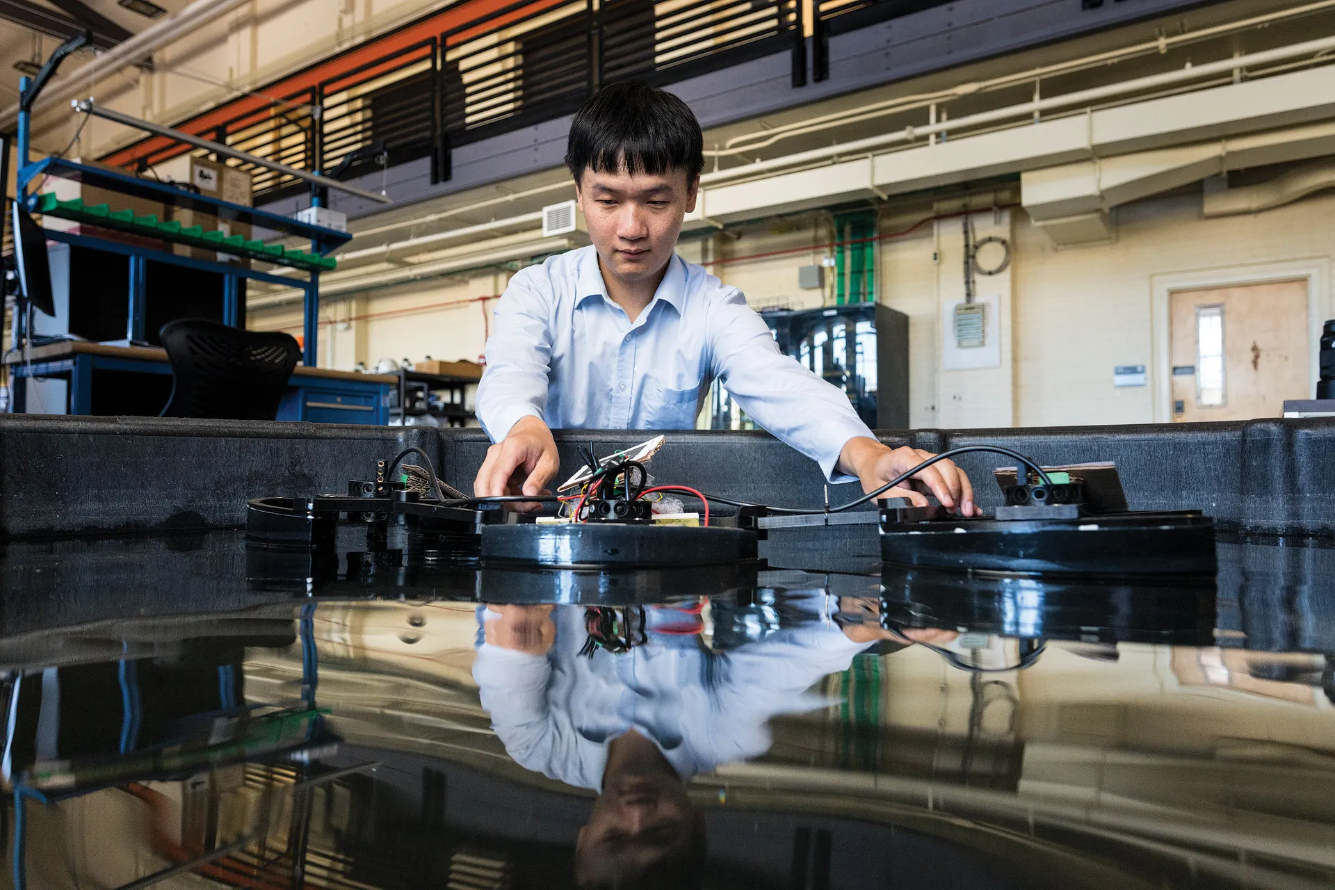 A graduate student reaching over a large container of water to adjust two underwater robots in a testing facility. 