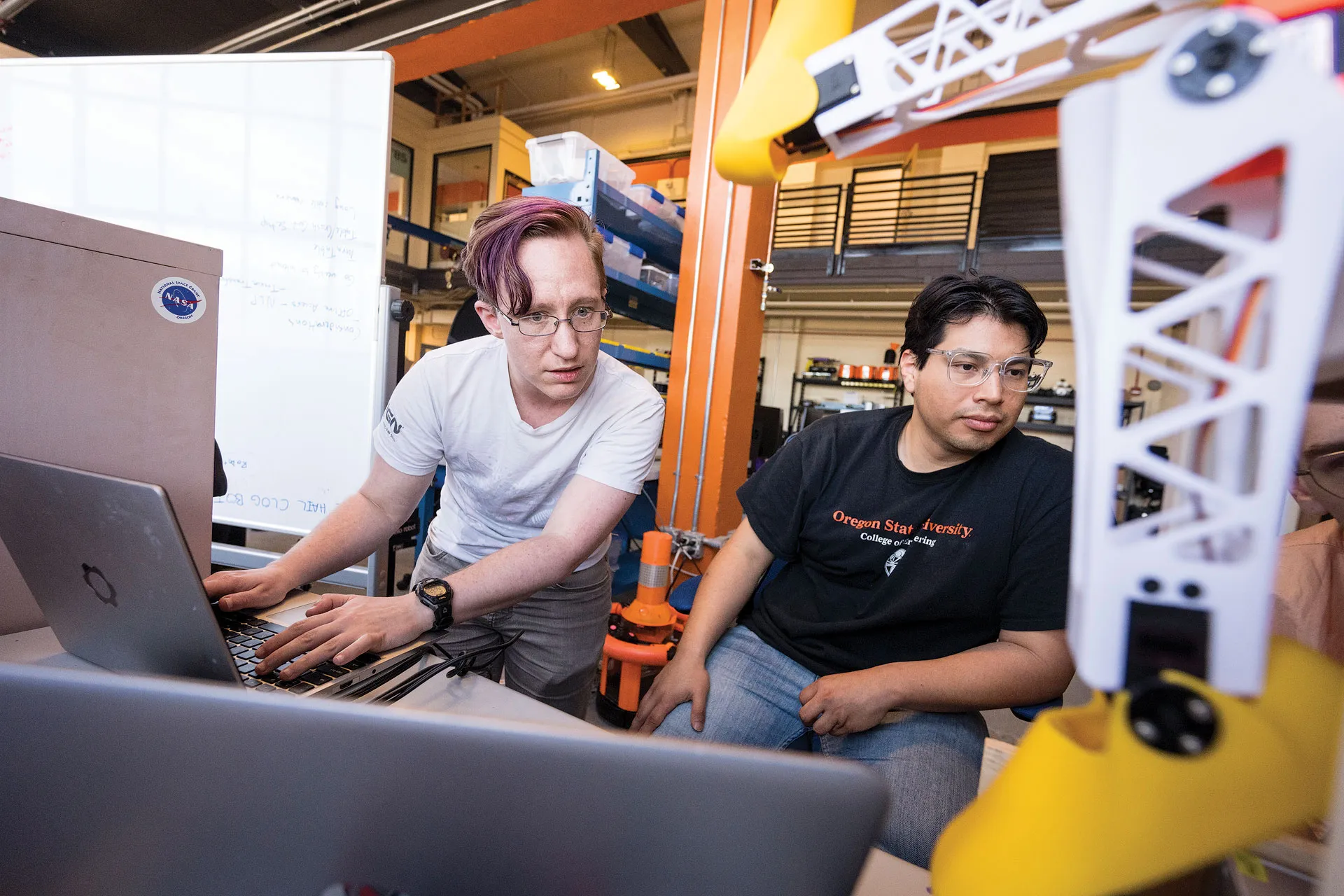 Two graduate students sitting at a desk in a large research facility looking over their laptop at a robotic shoe.