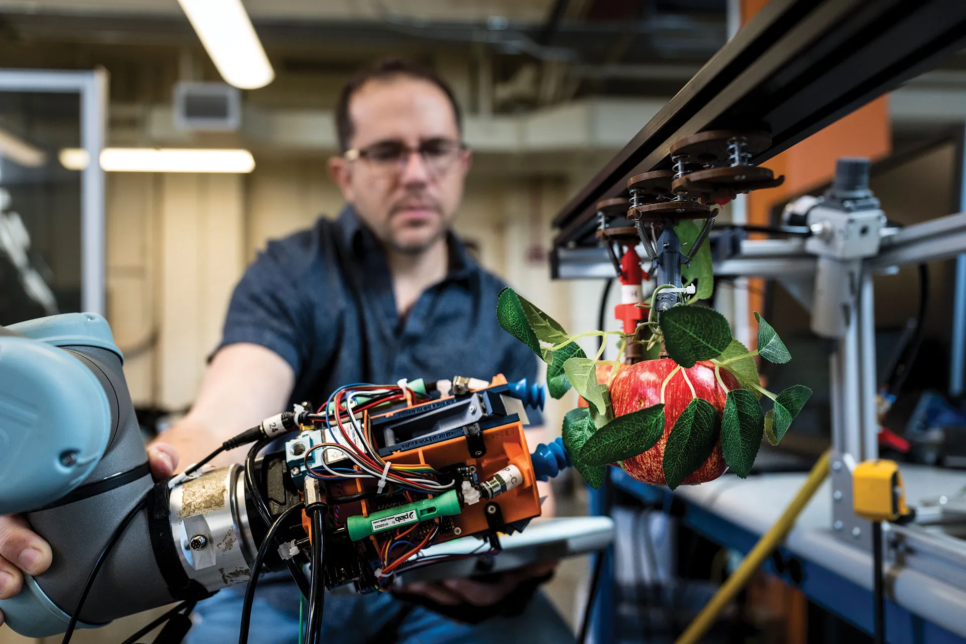 A robotic arm picking an apple in a research facility with a graduate student in the background adjusting the robotic arm.