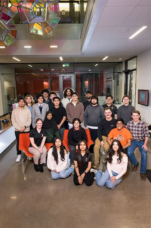 A group of 21 students sit and stand in a group within a lobby of campus building with a glass sculpture suspended above them.