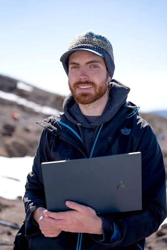 Ian Rankin, a person wearing a blue beanie, hoodie, and jacket, holds a laptop while standing on the side of a mountain, with a patch of snow and clear blue sky behind them.