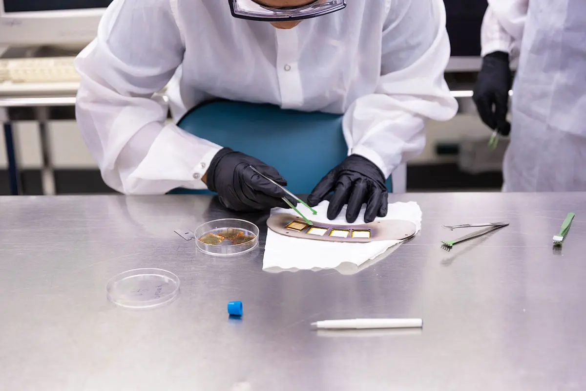 A person in a cleanroom suit bends over a lab table and uses tweezers to move to move materials.