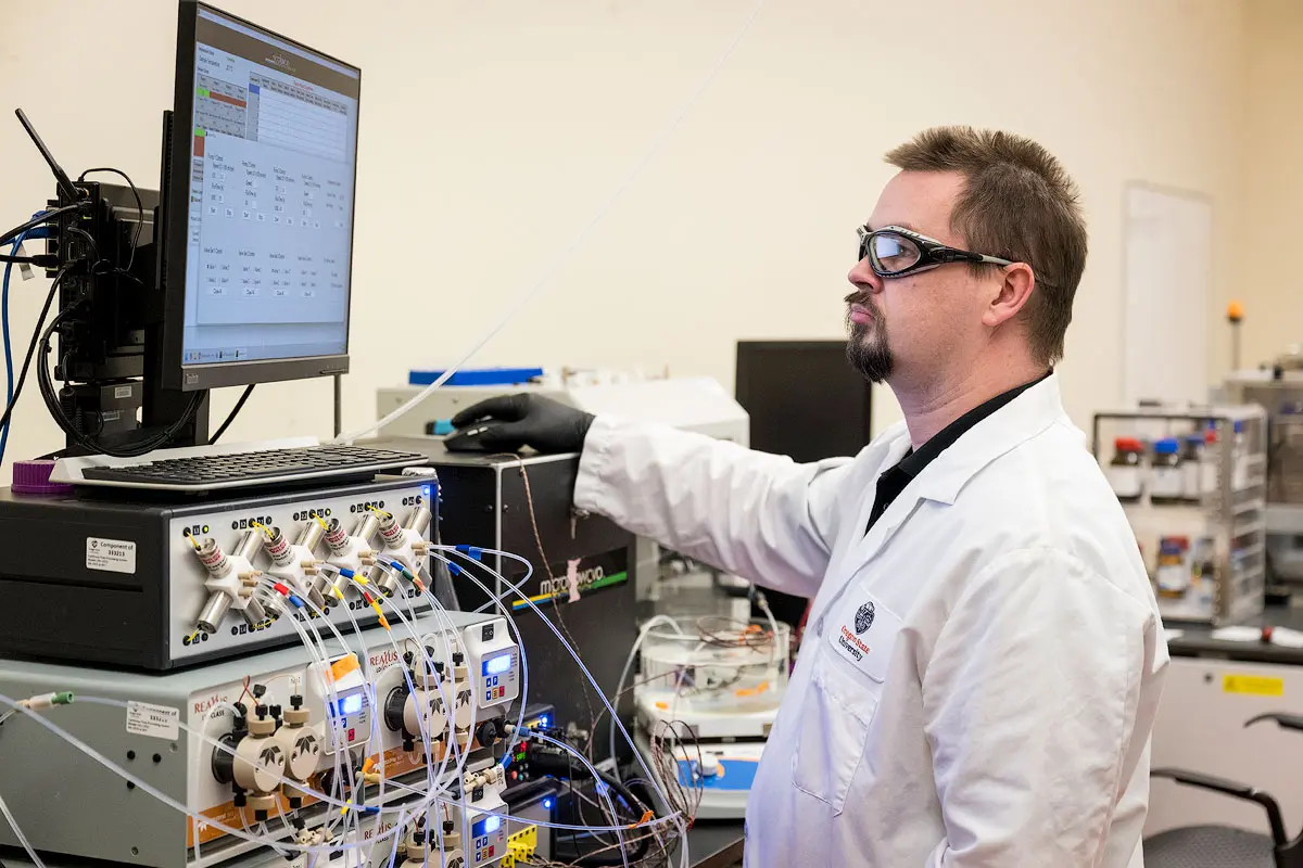 A person wears personal protective equipment in a lab and stands in front of equipment and a computer using the mouse.