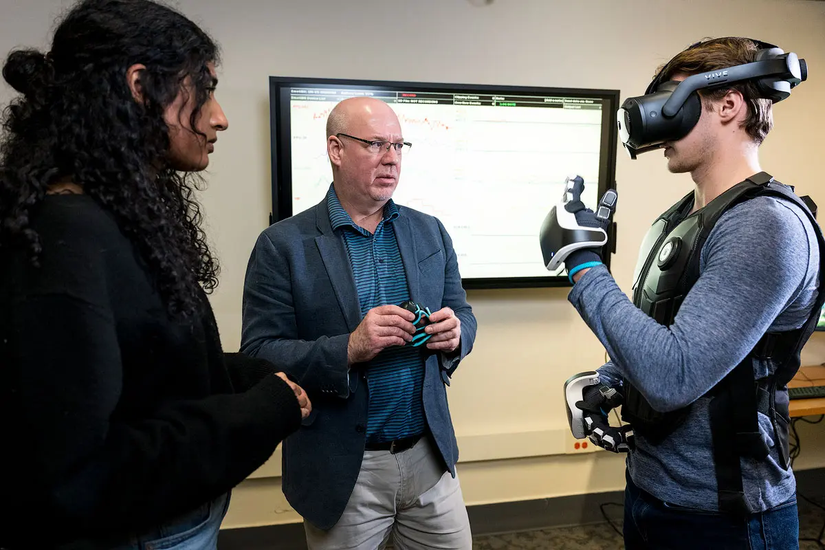 Three people stand in a lab with a digital screen in the background. One person wears an augmented reality headset and gloves.