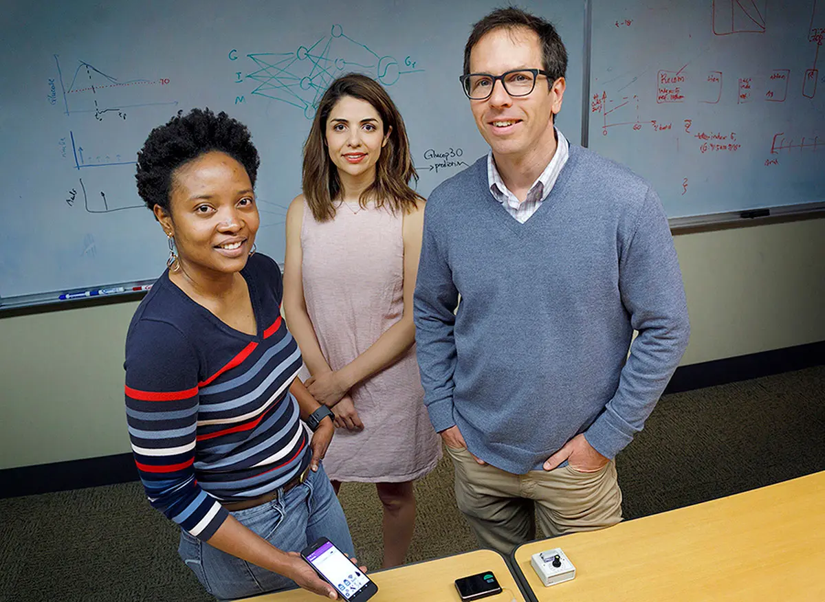 Three people stand in a classroom in front of a whiteboard with one person holding a smartphone in their hand.