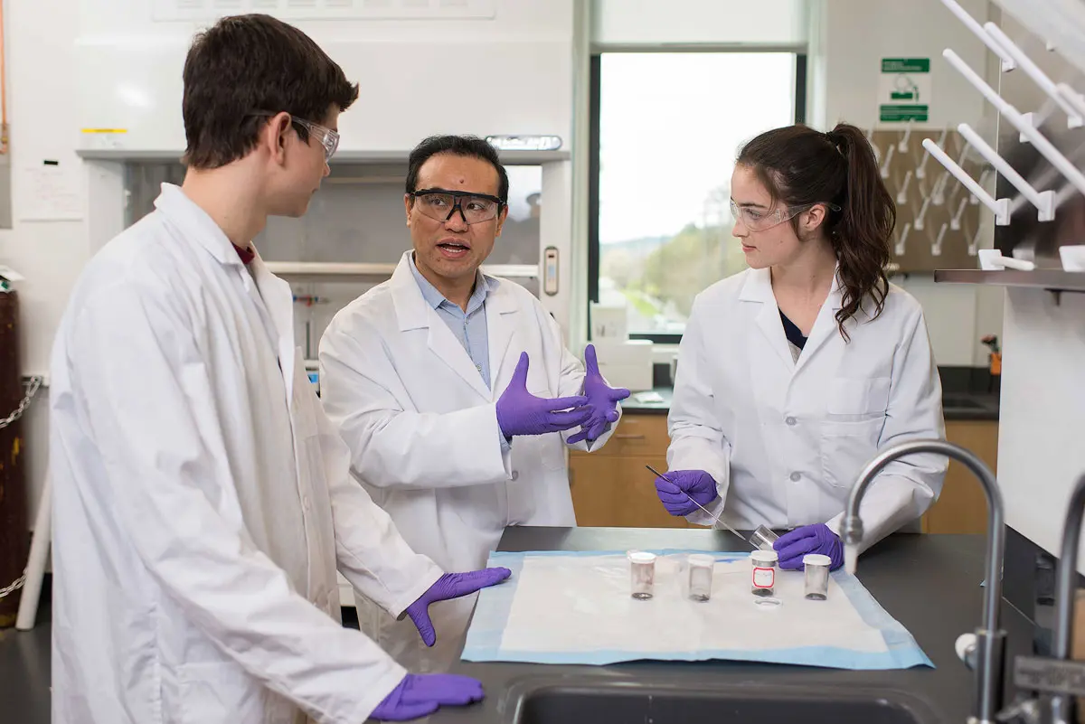 Three researchers in lab coats and eye protection stand over a table with five samples in containers.
