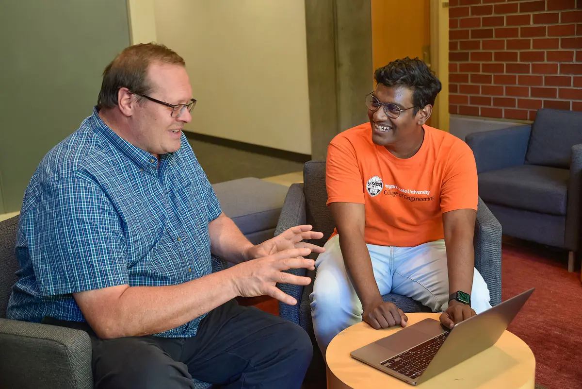 Two people sit in chairs and talk with a laptop in front of them. 