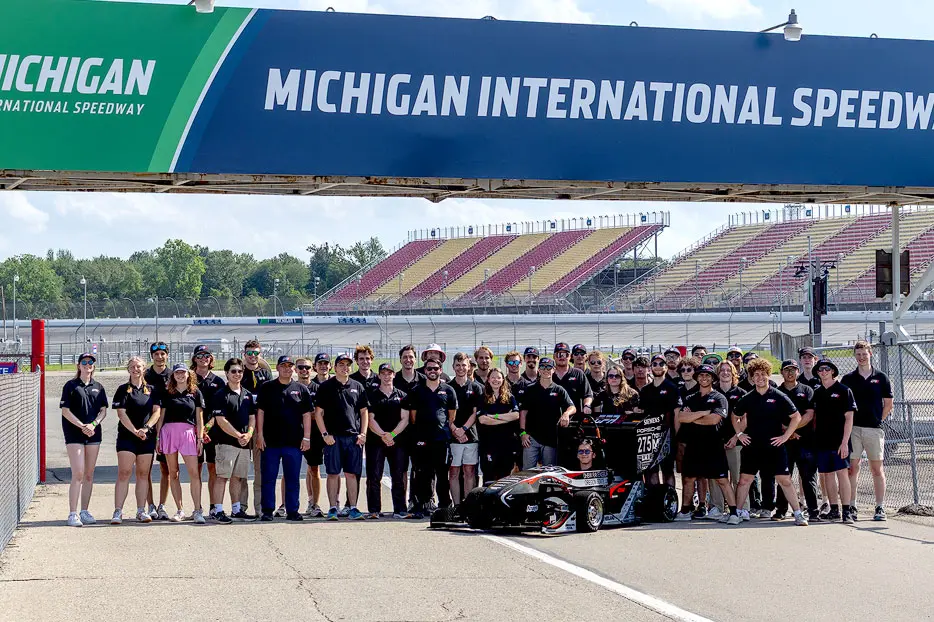 A group of about 40 people stand in matching black shirts on a track and face the camera with a race car in front of them.