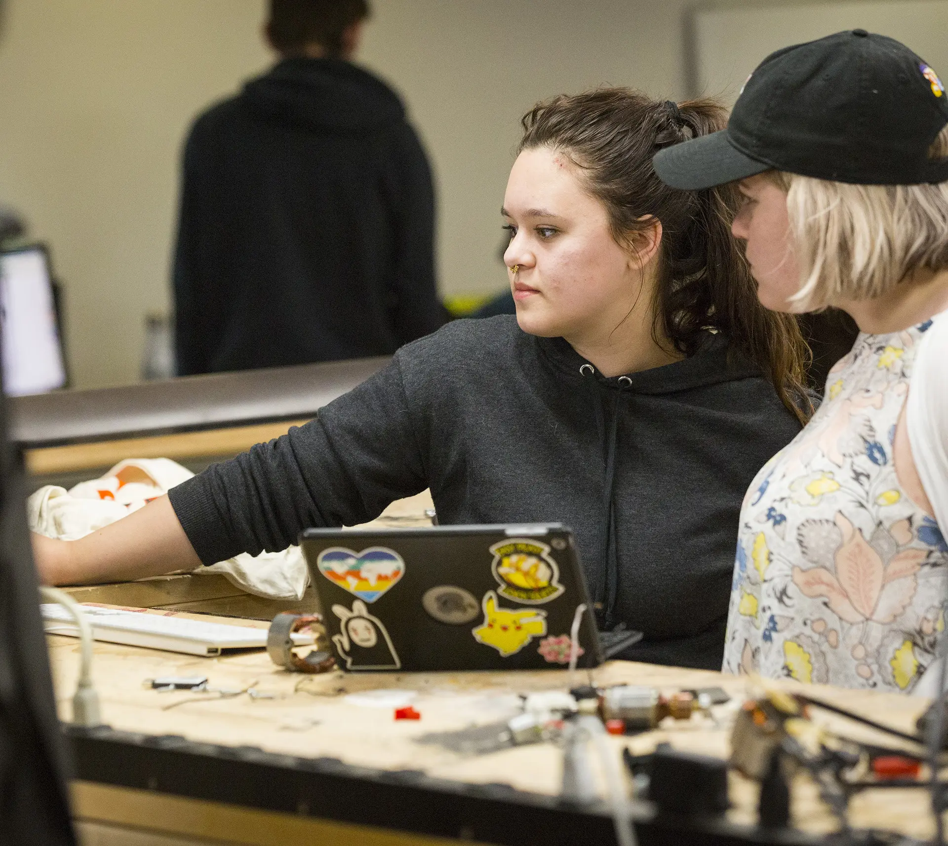 Two people collaborate at a workbench, using a sticker-covered laptop and electronic components in a lab.
