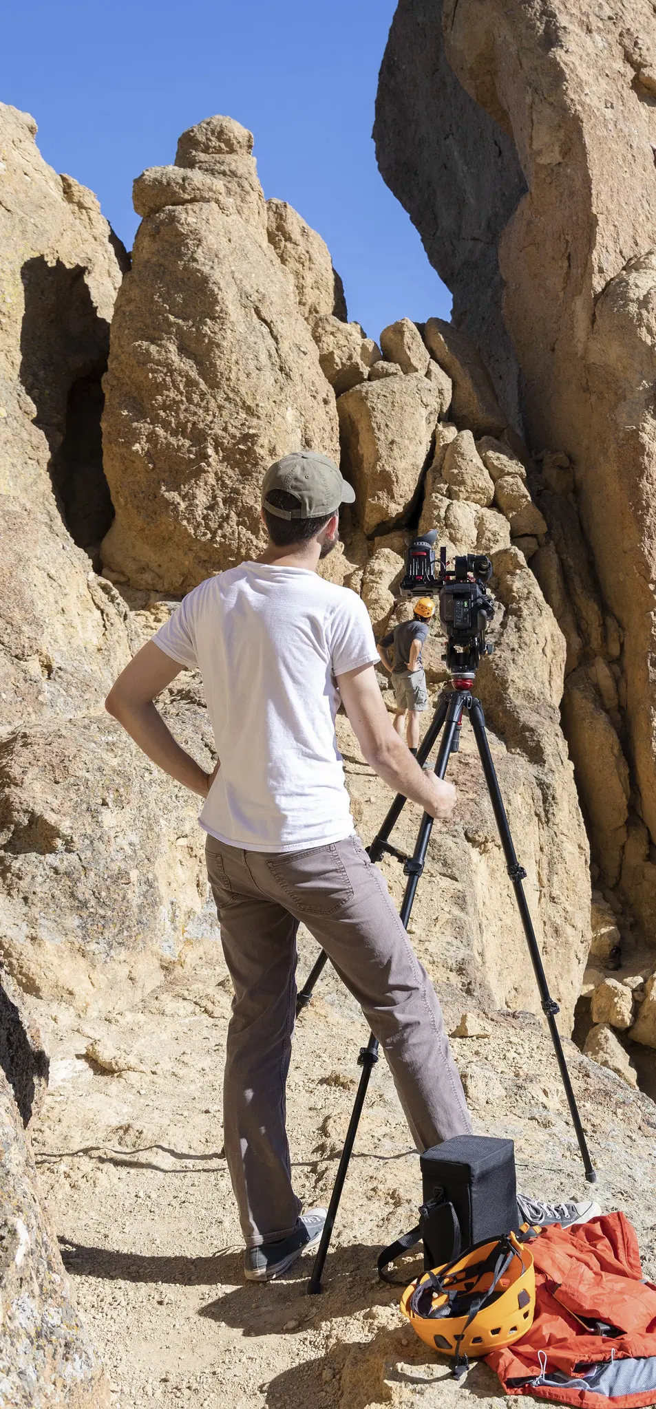 Person operating a camera on a tripod in a rocky area, with another individual climbing in the background.