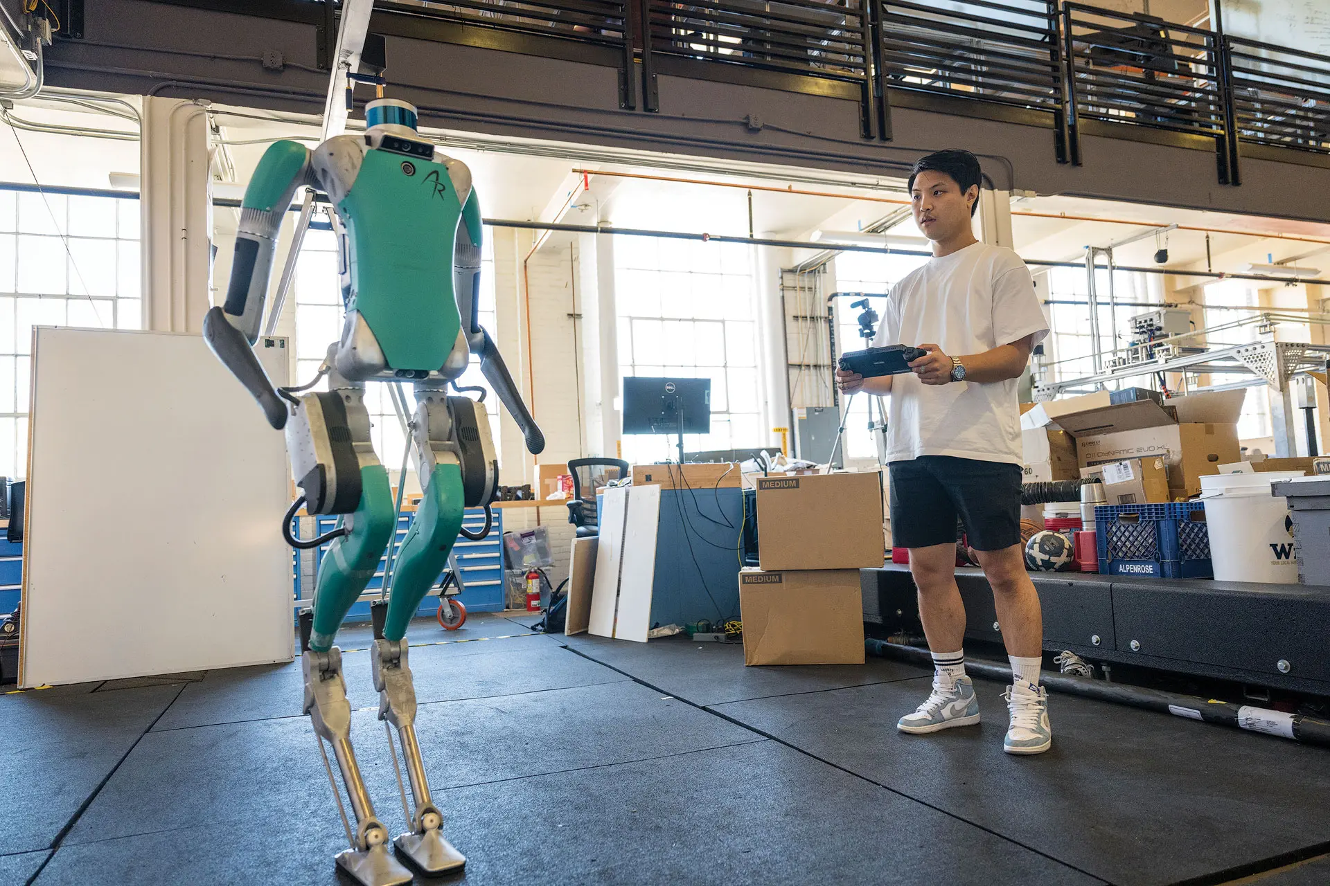 A graduate student standing in a large facility with a controller in his hands with Digit, the bipedal robot with long arms in the foreground.