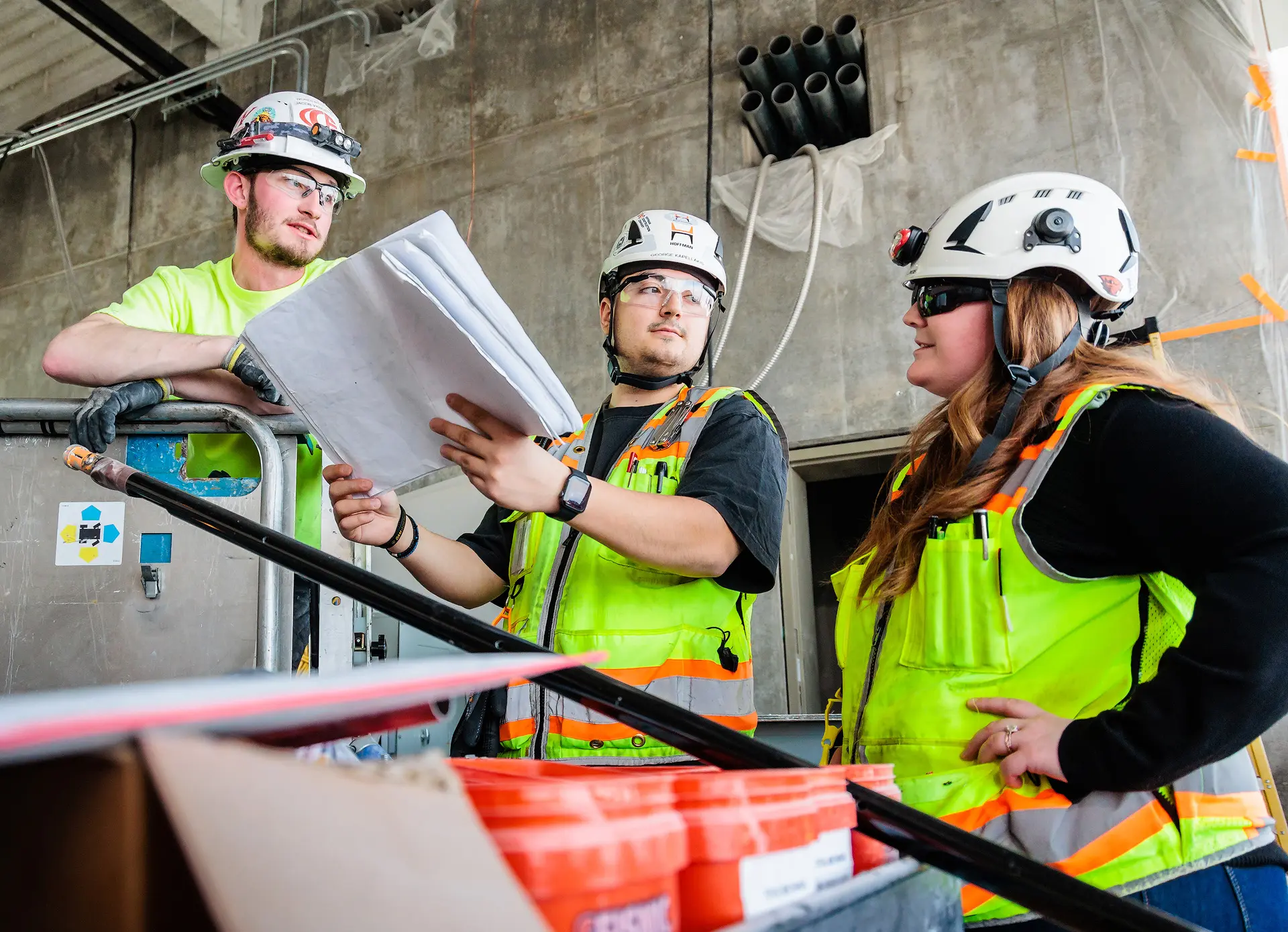 Three construction workers in hard hats and safety vests review plans in an industrial setting.