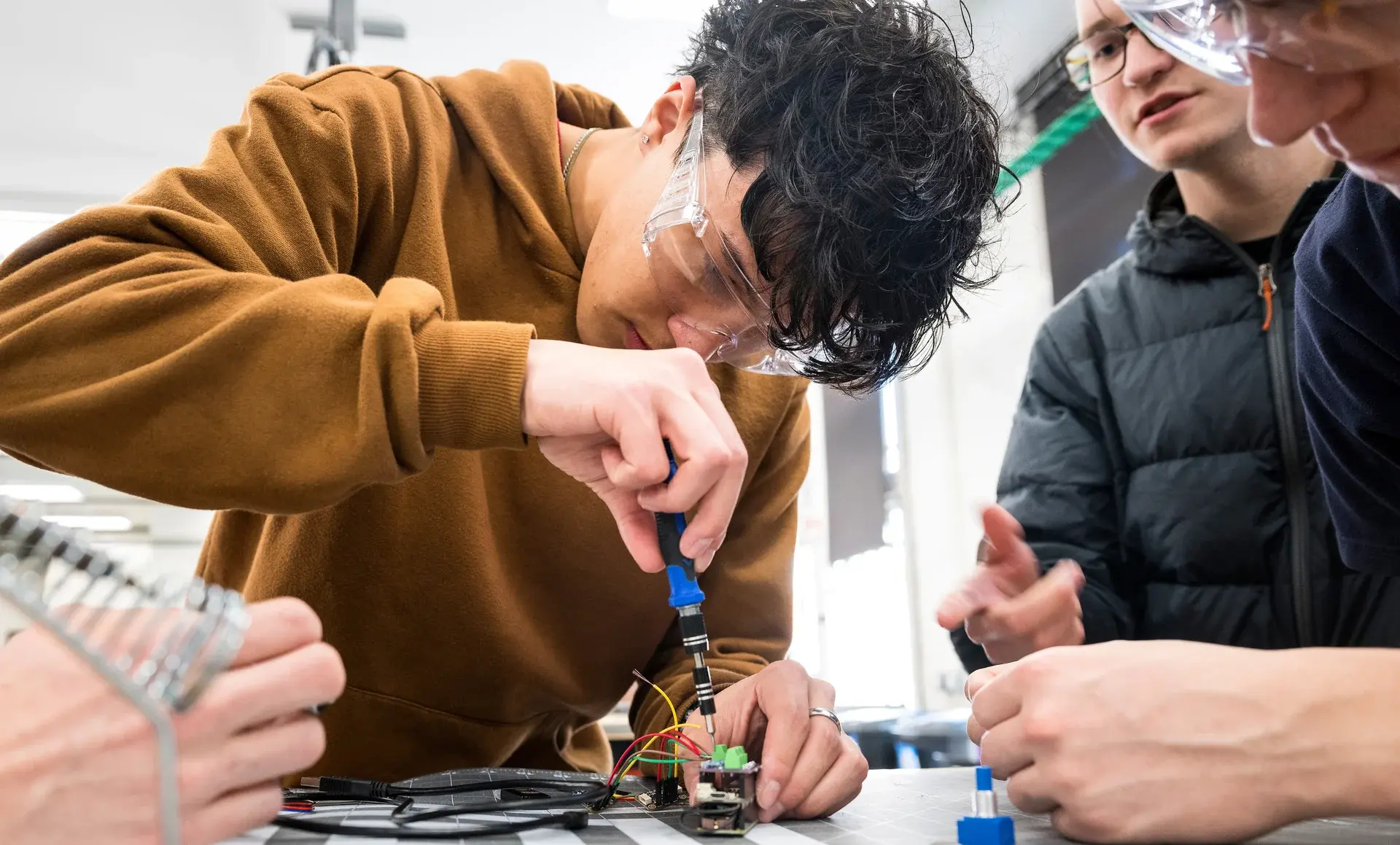 Student using a screwdriver on a circuit board while classmates assist at a workbench.