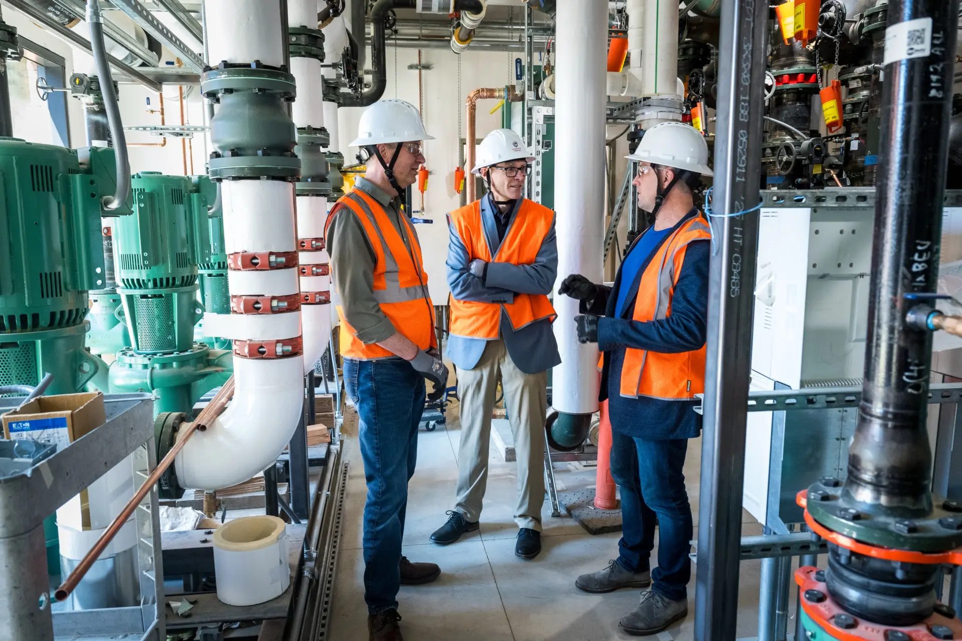 Koppers, Palmer, and Huddleston wearing hard hats and safety vests surrounded by a variety of large pipes.