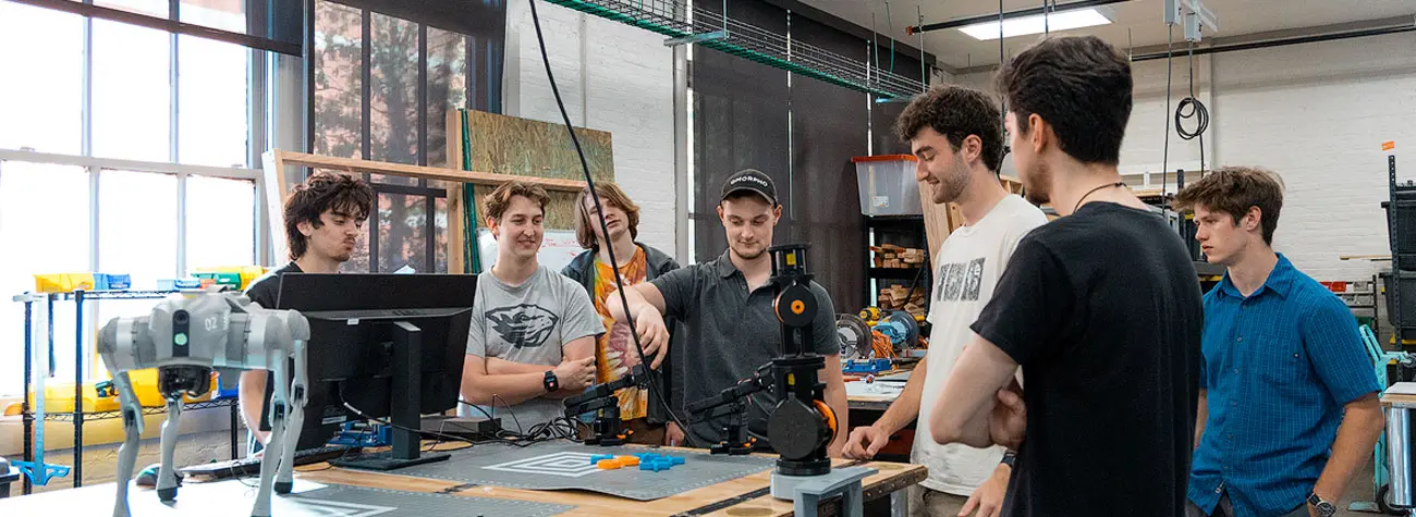 Seven students gather around a workbench while one manipulates a robotic arm.
