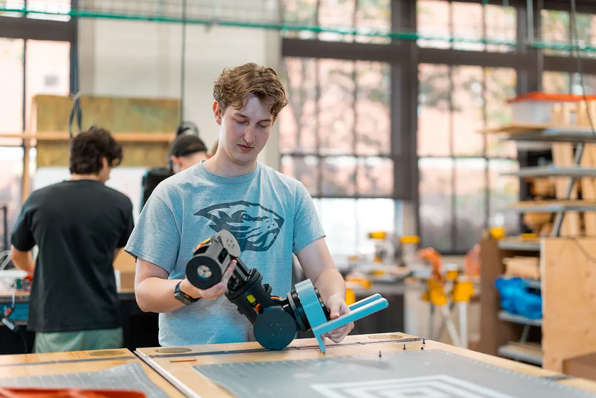 Alexander Roller wears a grey Oregon State Beavers t-shirt and stands in a workspace and examines a small robot arm over a bench.
