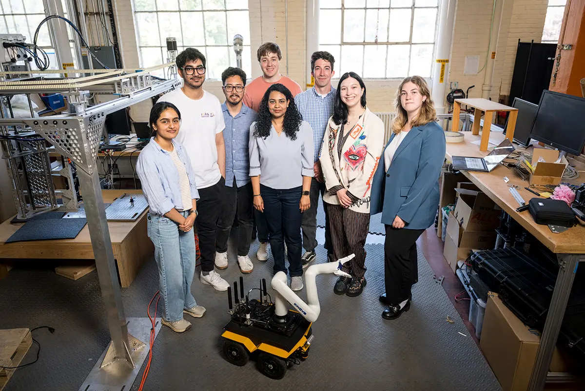 Eight people stand with Sandhya Saisubramanian in a lab and look toward the camera with a robot with an arm on the ground in front of them. 
