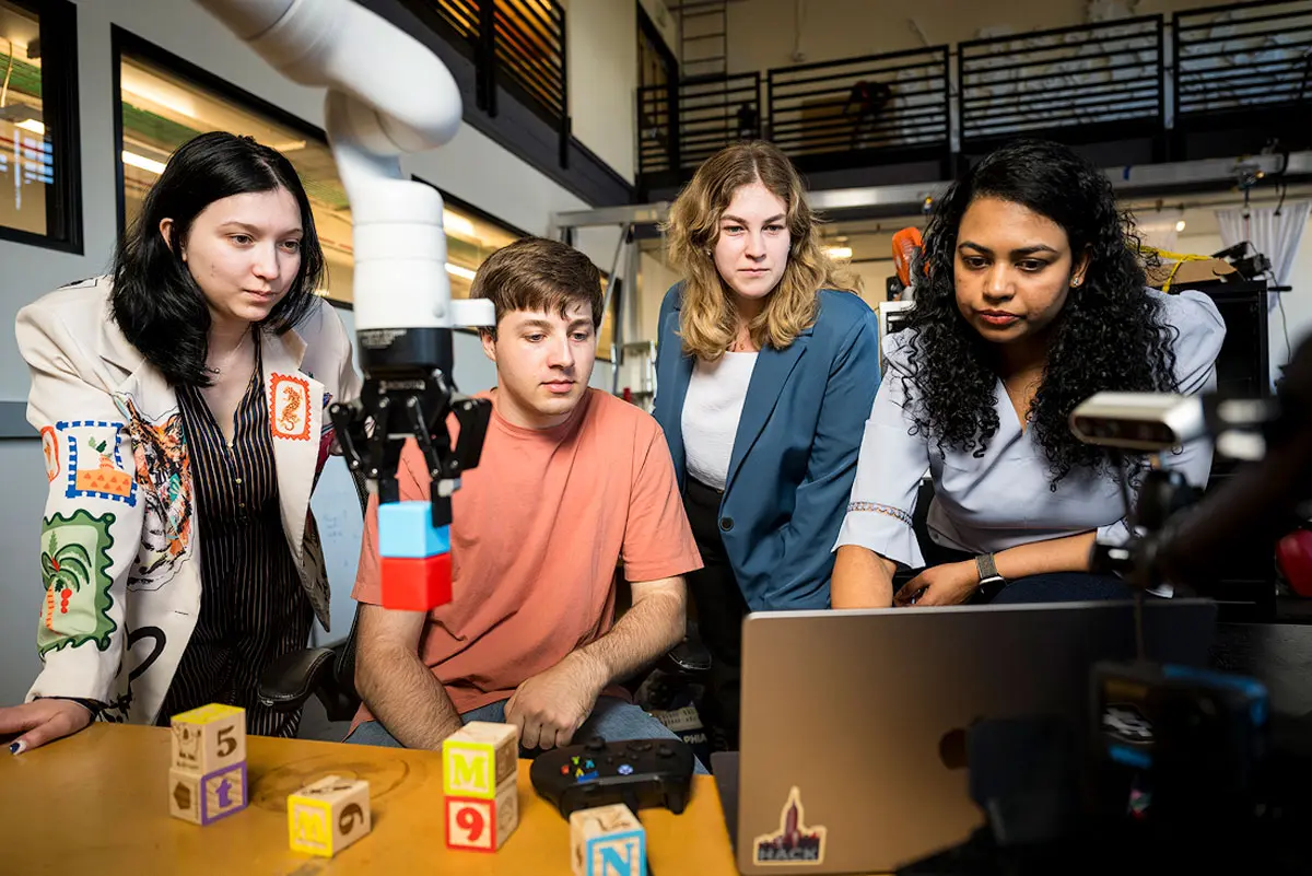 Three people stand next to Sandhya Saisubramanian and lean over a workbench as a robotic arm picks up children’s block toys and they look at a laptop in front of them. 