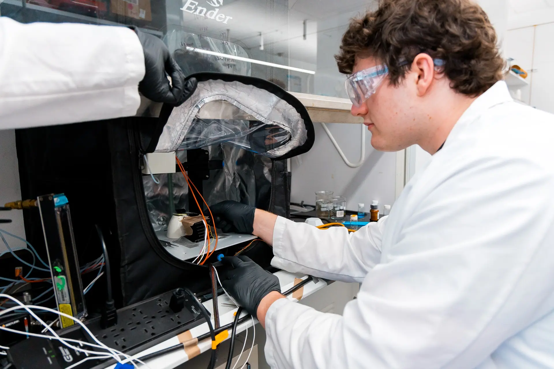 tudent Anton Escher, where white lab coat, goggles, and black gloves manipulated a cylindrical device under a chemical hood.