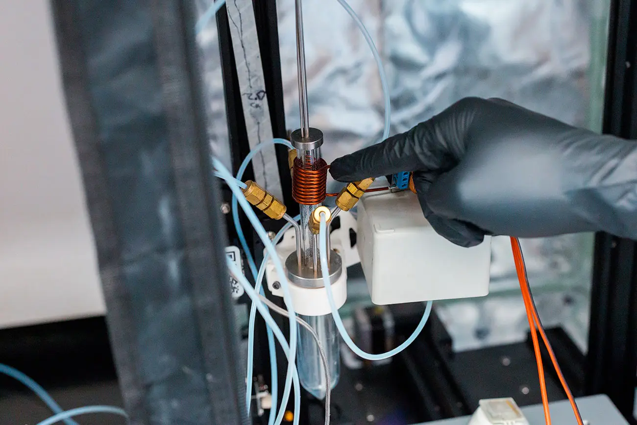 A gloved hand points to a lab device with tubing and a copper coil around a small cylinder.