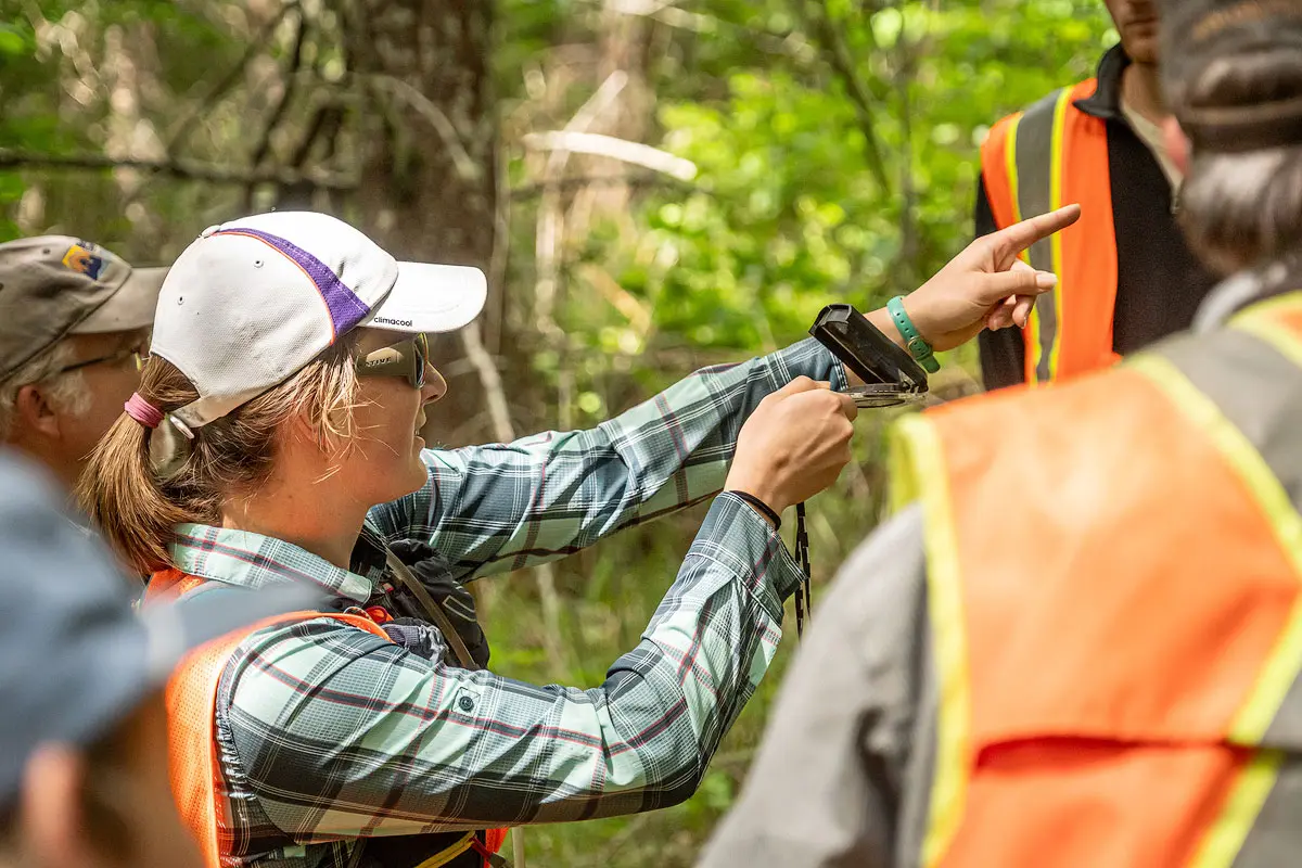 A person in an evergreen forest holds a compass and directs a group of students to the designated direction.
