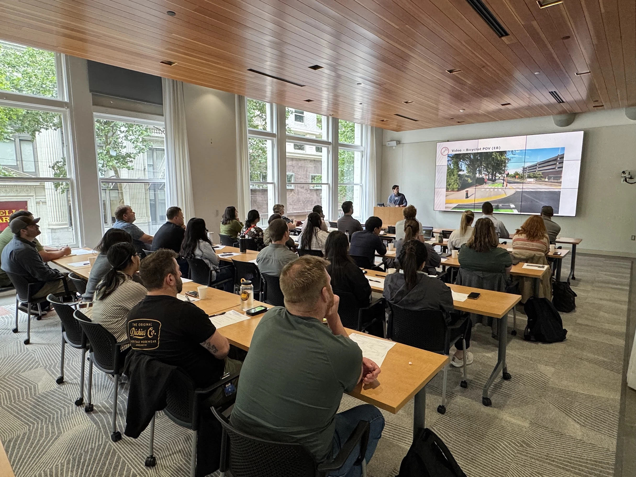 A presenter addresses a seated audience in a bright classroom while a screen shows a bicyclist’s view of a street.