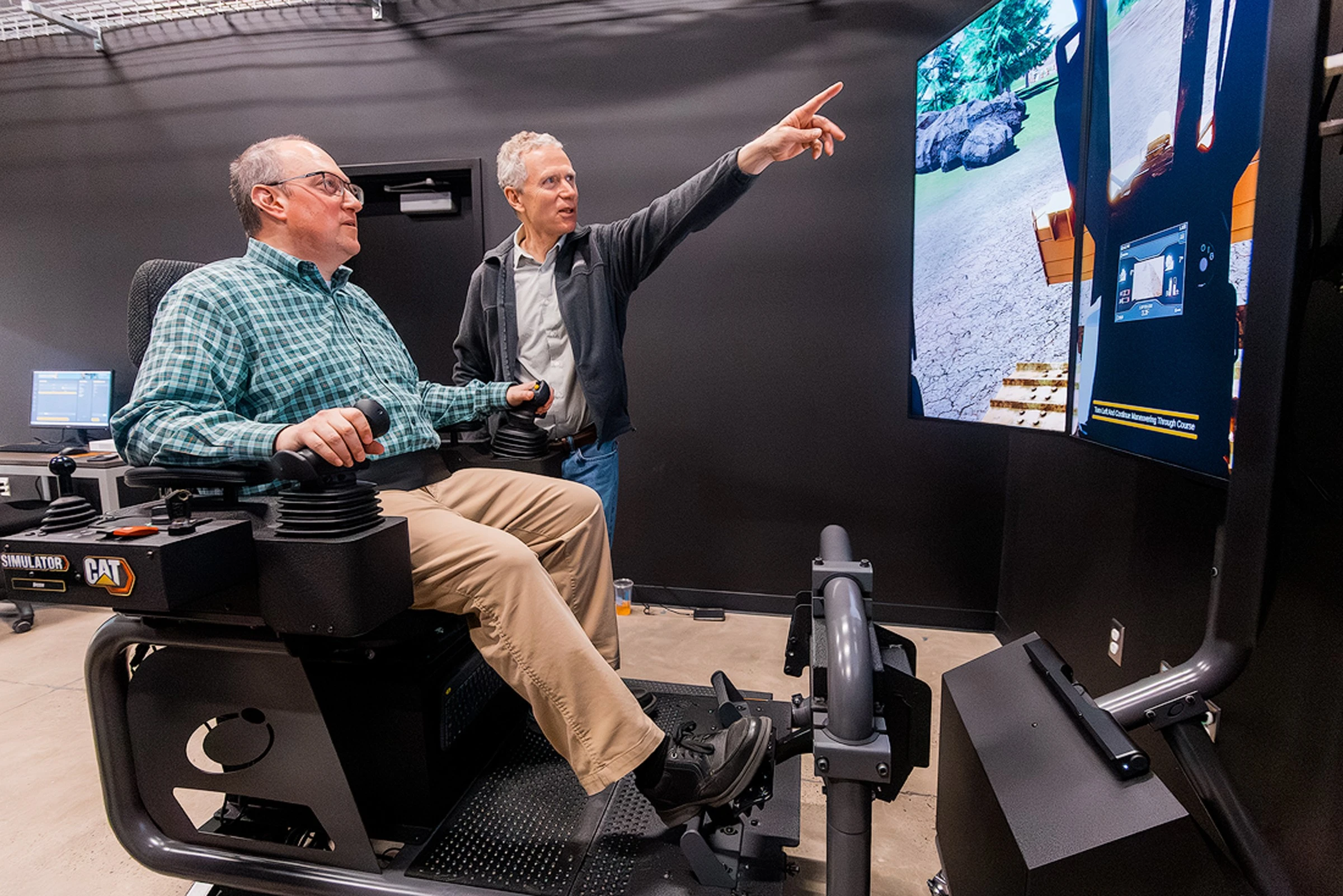 David Hurwitz operates a CAT heavy equipment simulator while another person points to a display showing a virtual construction site.