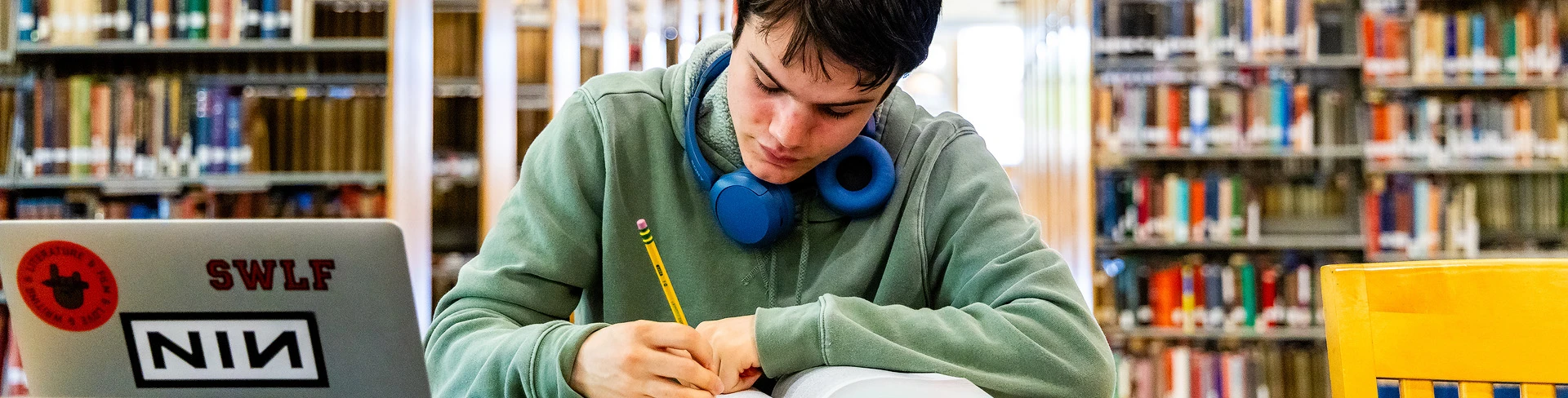 A student writes with a pencil at a table in the library.