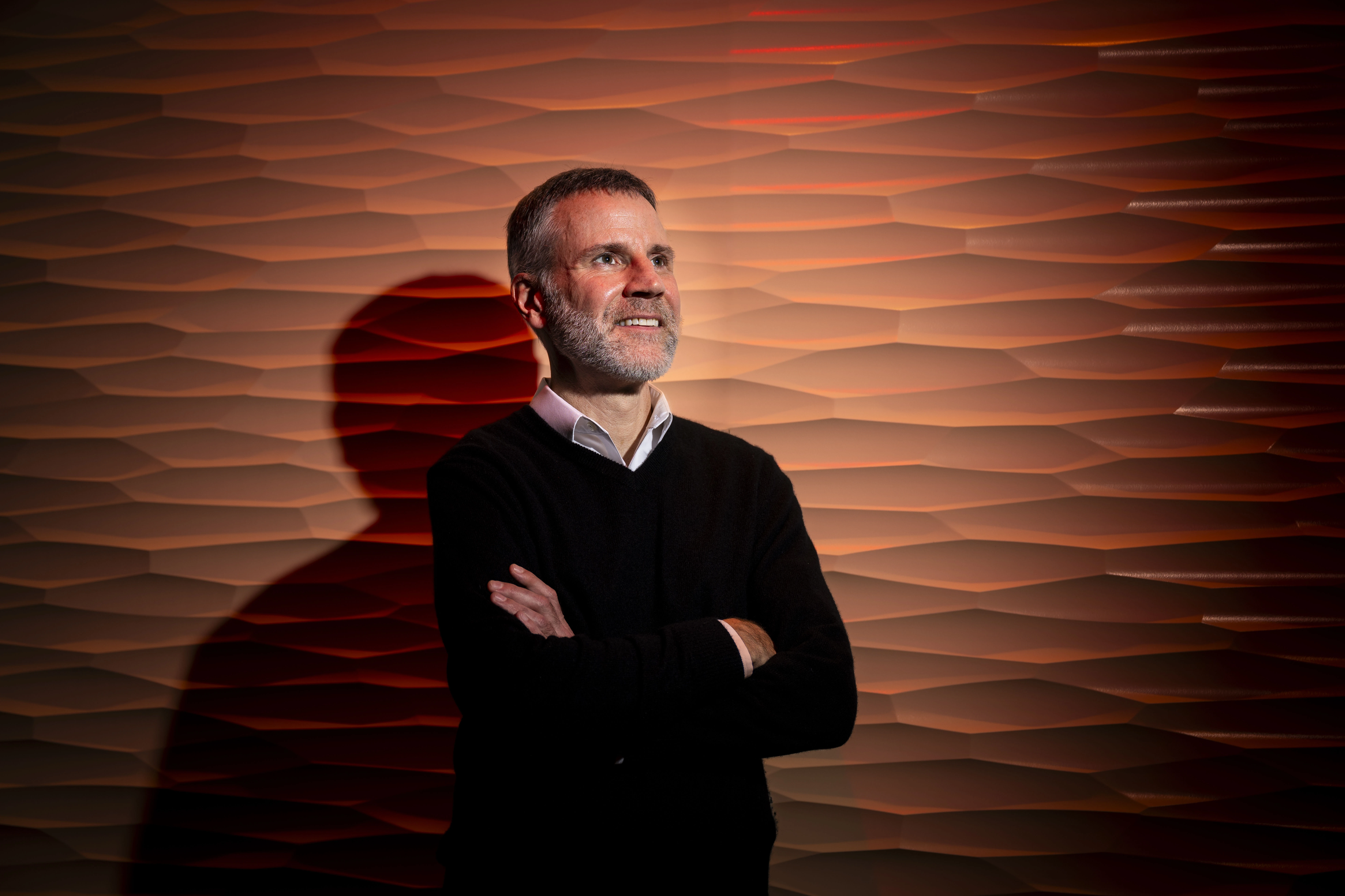 Kevin Houser stands with arms crossed before a glowing, layered orange wall, casting a long shadow in warm light inside Oregon State University’s Lighting Lab.