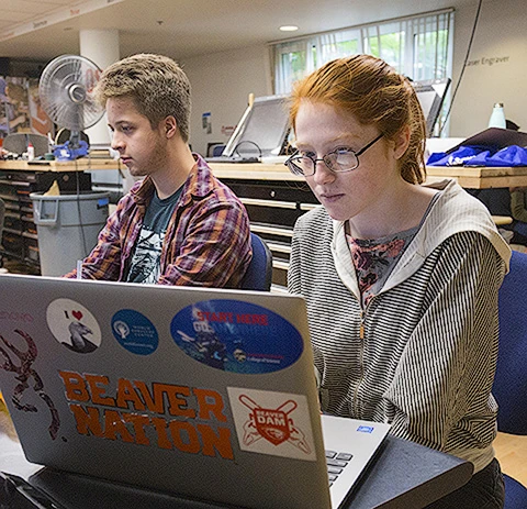 Two students working at a desk on their laptops.