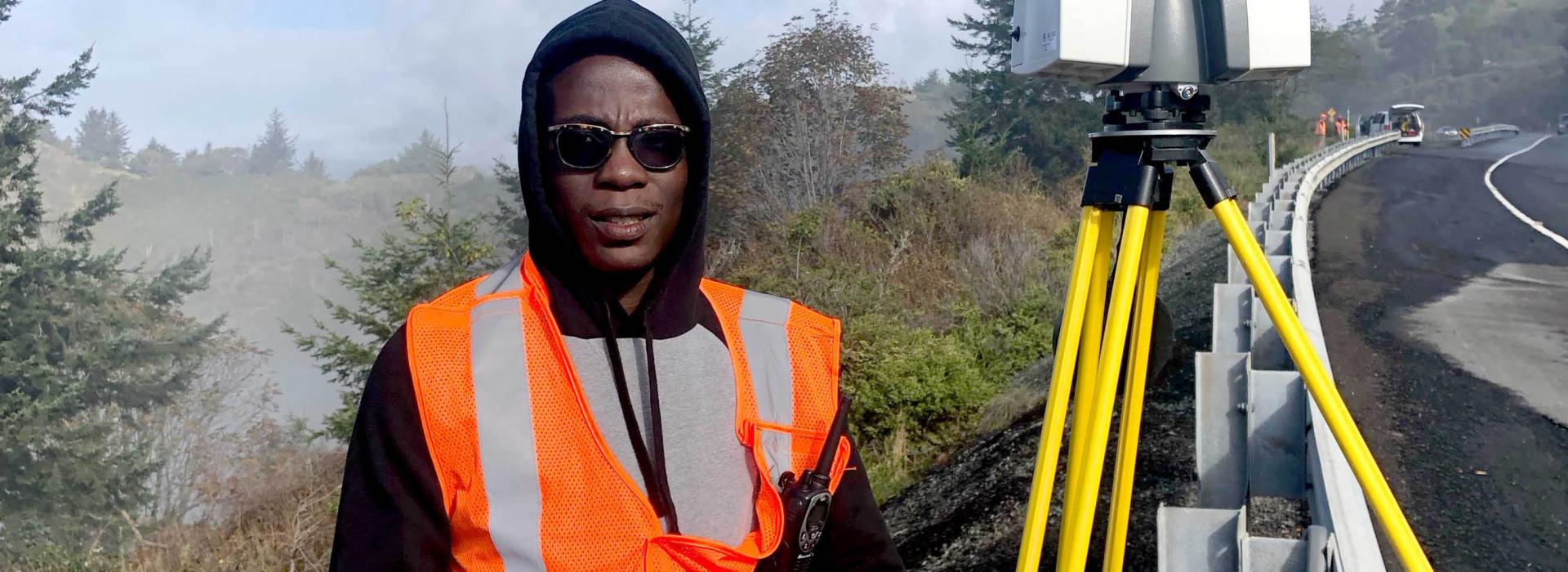 A student standing on the side of a coastal highway next to surveying equipment.