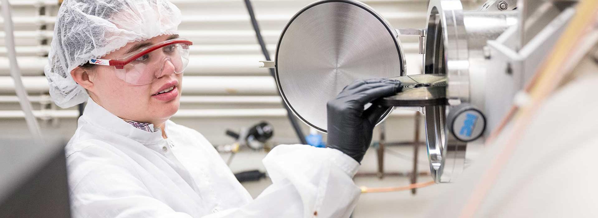 A graduate student in personal protective equipment removing a sample from equipment in a cleanroom.