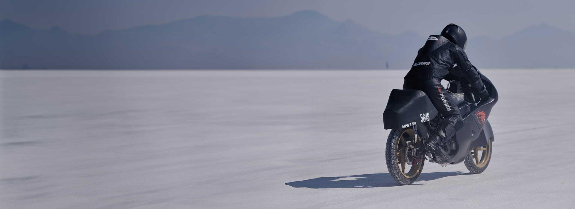 A motorcyclist riding a 50cc motorcycle across desert sand with mountains in the distance.