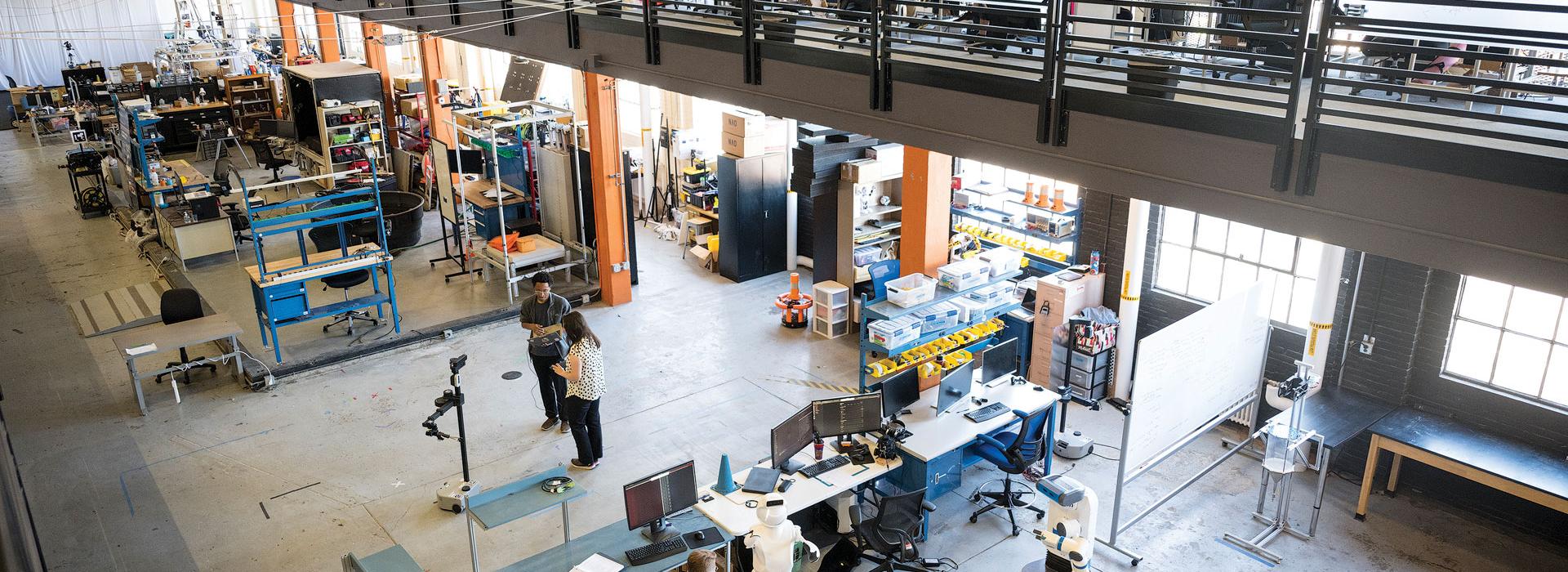 Two researchers standing in the center of a large open-room, multi-story research facility with robotics equipment and desks surrounding them. 