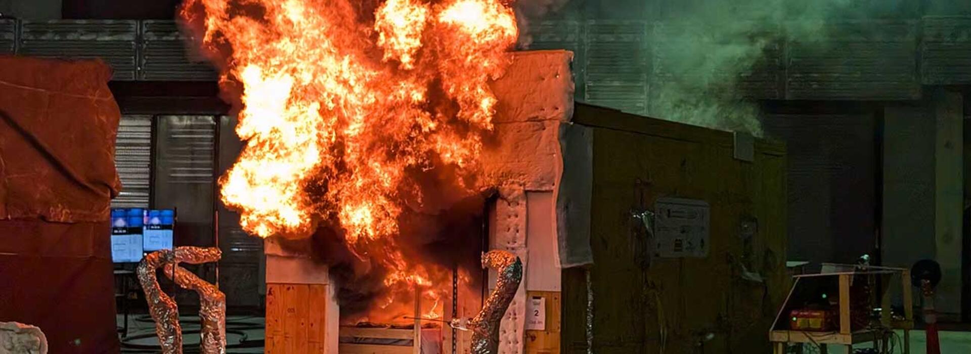 Fire emerging from a small structure within a larger testing facility. 