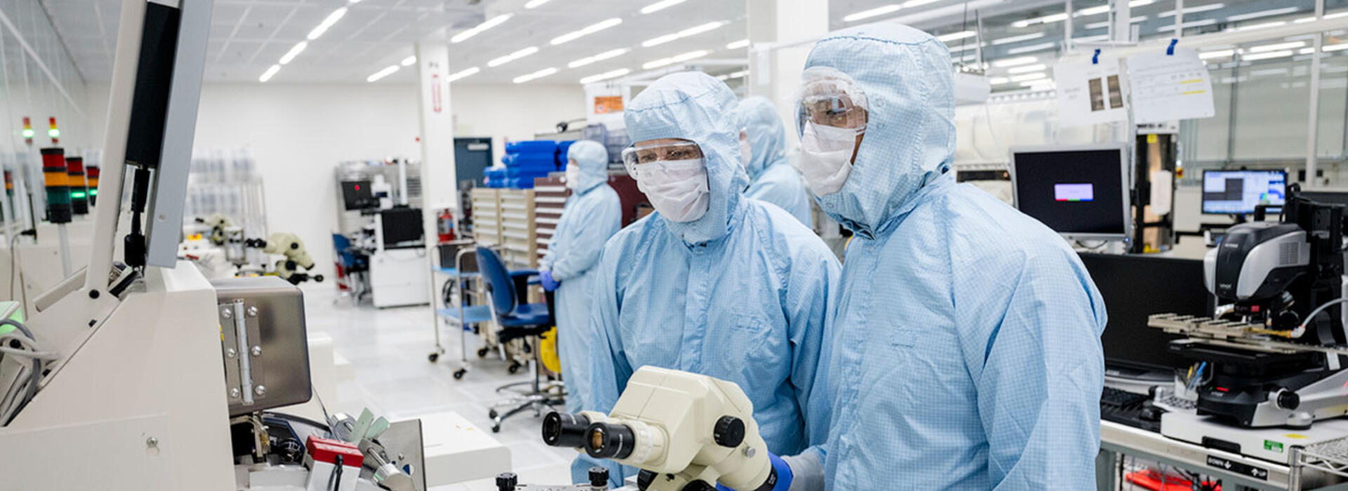 Two people wear cleanroom suits and stand in front of a microscope. 