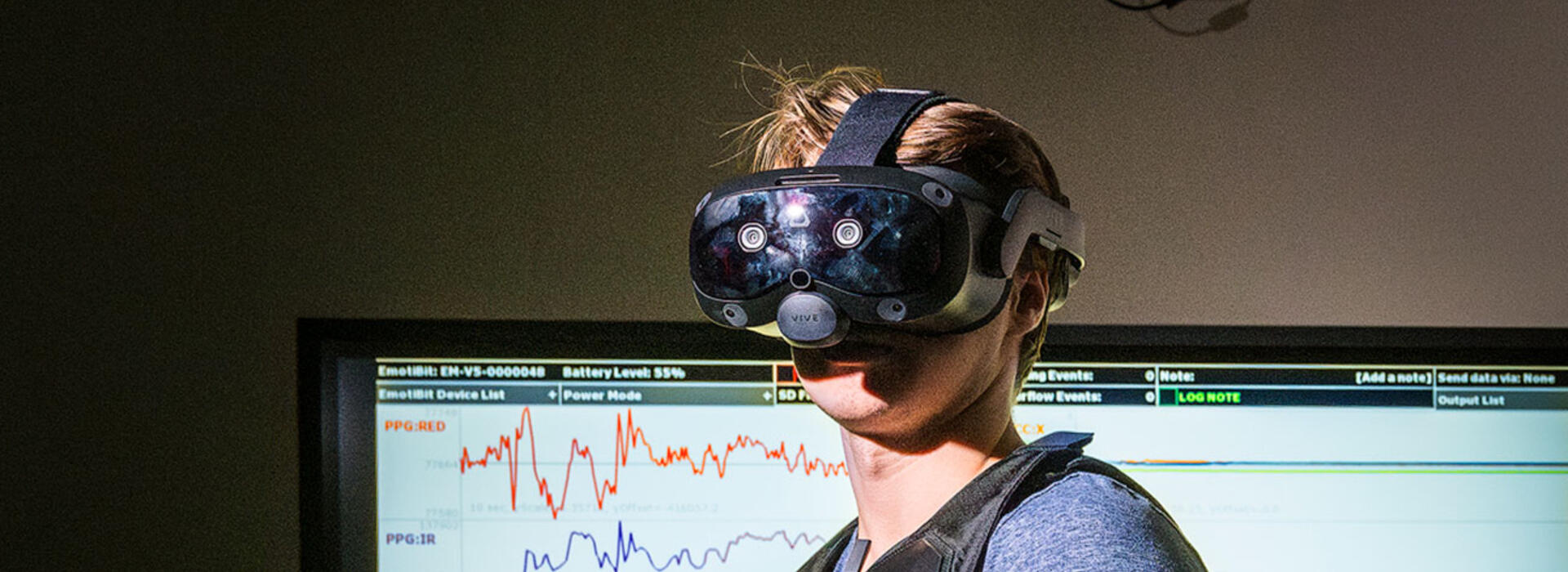 A person wears an augmented reality headset inside a lab and a digital screen displays data in the background.