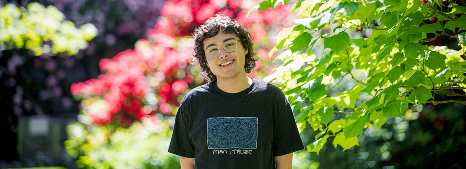 A person wearing a t-shirt smiles at the camera and stands in front of trees and spring blooms.