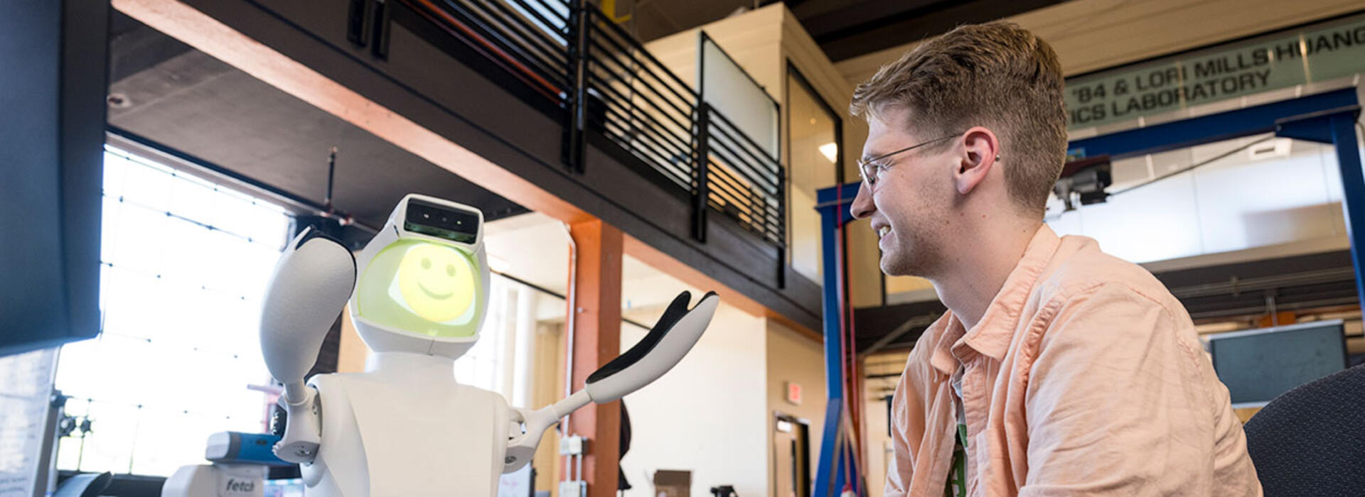 A person sits in a lab and looks at a robot with a smiling face and arms raised upward.