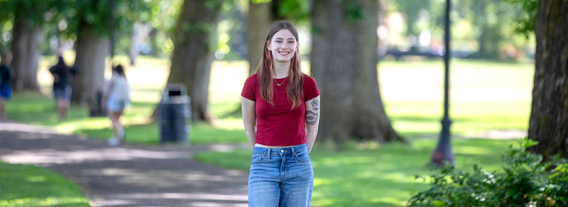 A person stands on a tree-lined path and smiles toward the camera. 