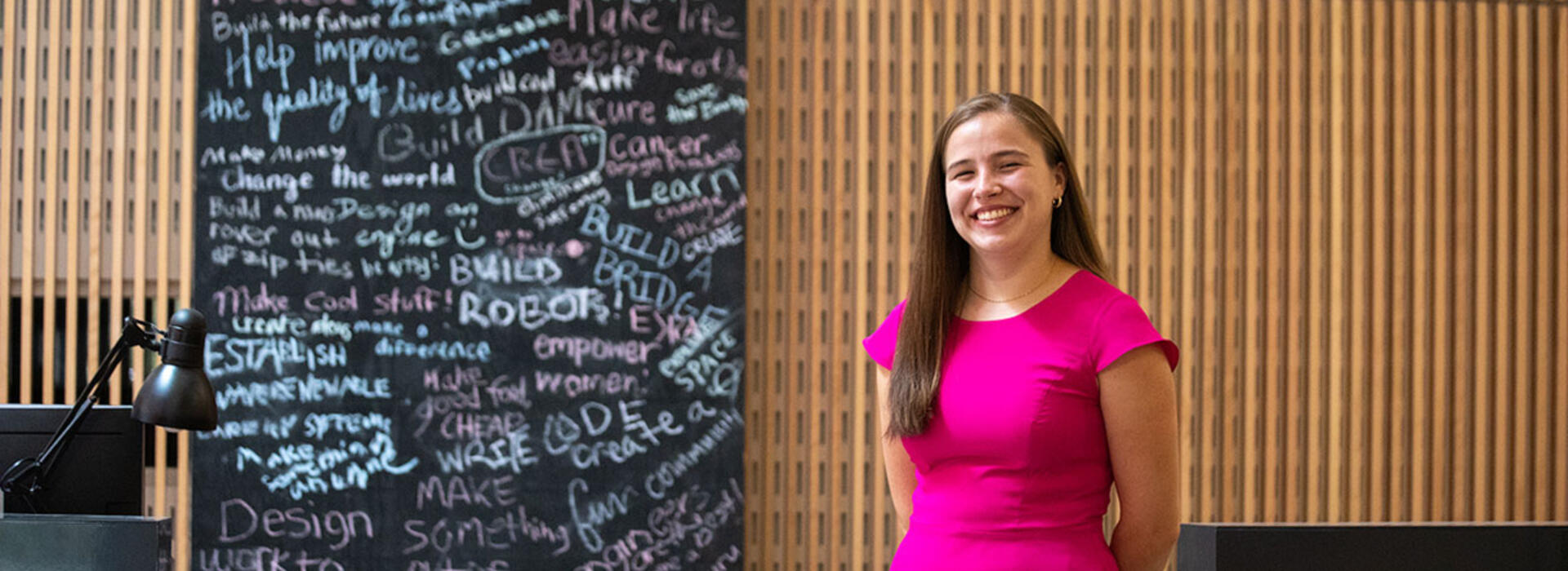 A person stands in front of a chalkboard with engineering terms written on it and smiles toward the camera.