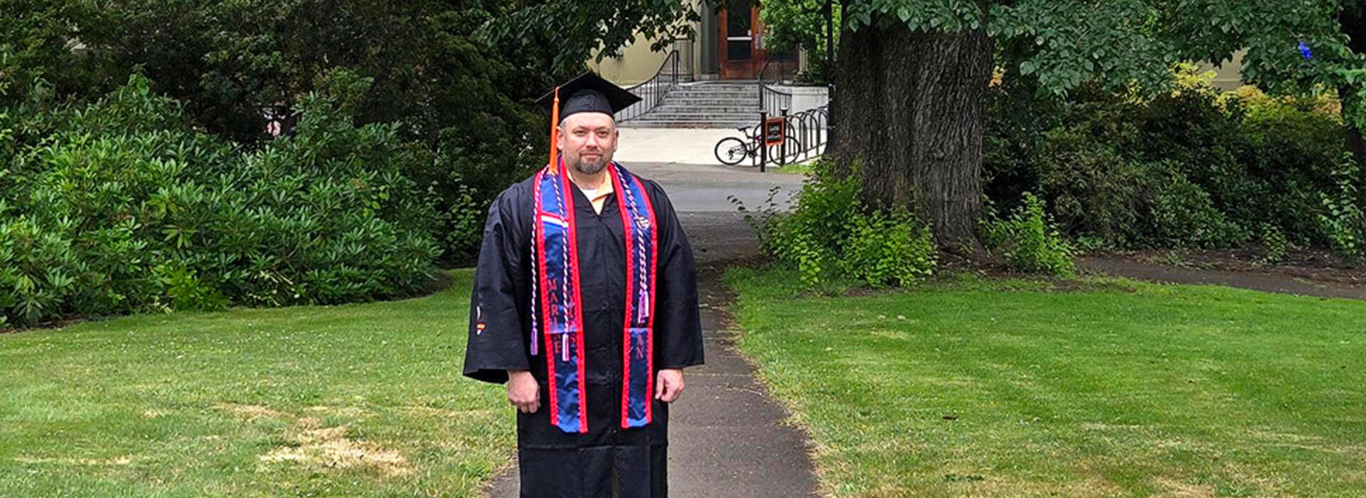 A person stands outside and looks toward the camera wearing academic regalia.