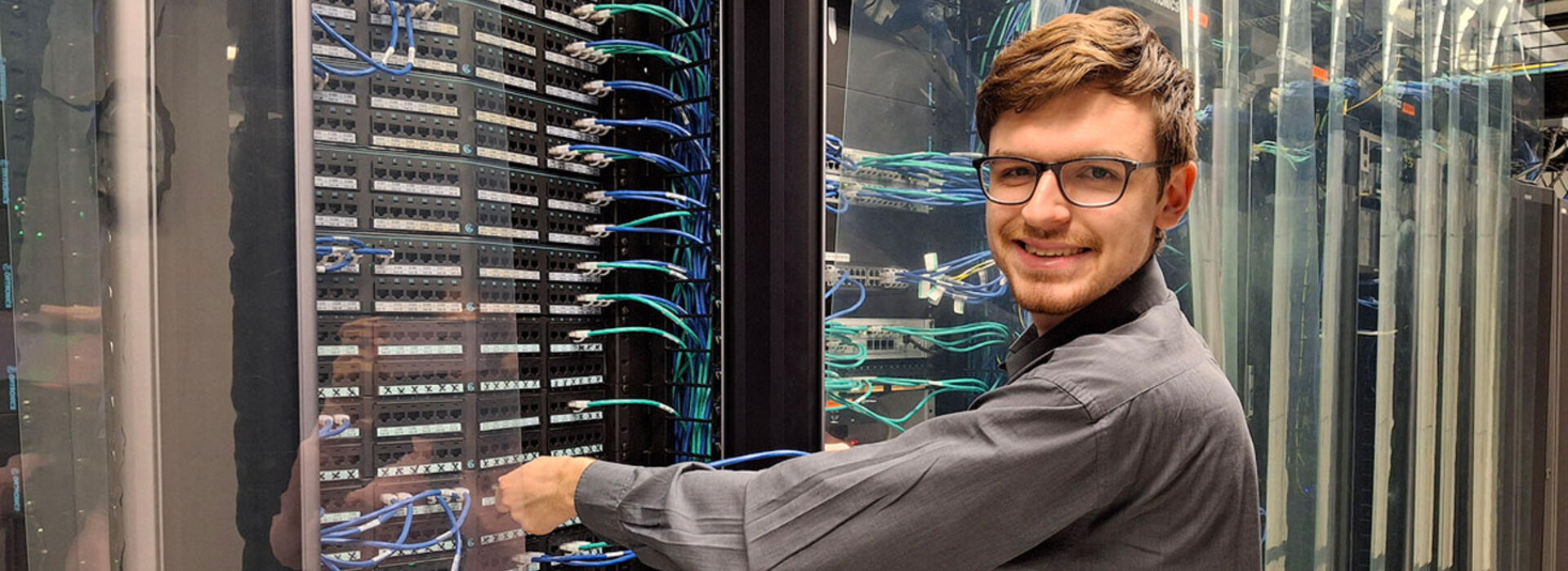 A person stands in a data center and adjusts hardware while looking toward the camera and smiling.