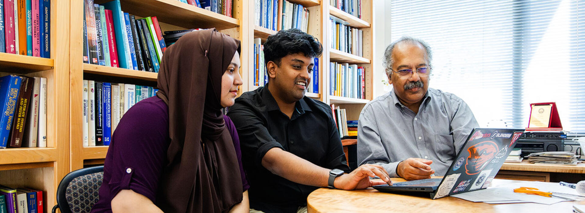Three people sit at a table in an office and look at a laptop. 